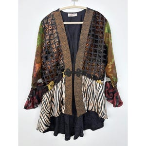 May include: A long-sleeved jacket featuring a mix of patterns including a black grid over leopard print, zebra stripes, and solid black panels. The garment has a brown patterned front border, a black frog closure, and ruffled sleeve cuffs.