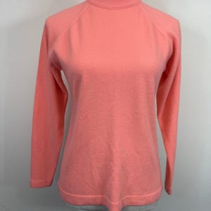 May include: A coral-colored sweater with a mock turtleneck and long sleeves. The sweater has a ribbed neckline and hem. The sleeves have a raglan design. The sweater is displayed on a mannequin.
