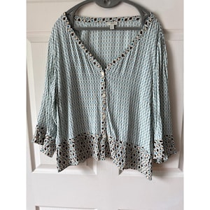 May include: A light blue button-down blouse featuring a repeating geometric pattern in dark brown and white. The garment has a V-neckline, three-quarter length sleeves with flared cuffs, and a contrasting patterned border along the hem and neckline.