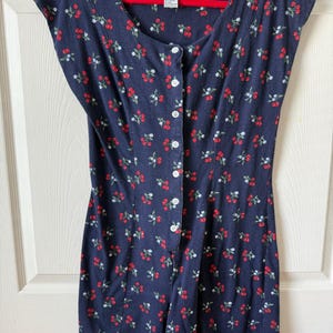 May include: A navy blue romper with a cherry blossom pattern. The romper has a button-up front, cap sleeves, and a scoop neckline. The brand name "energie" is visible on the tag. The romper is hanging on a red hanger.