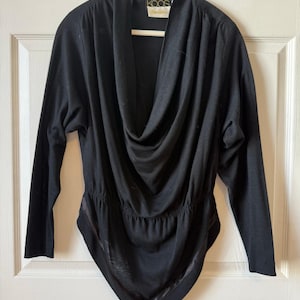 May include: A black long-sleeve top with a draped cowl neckline and a gathered waist. The bottom hem is a pointed, asymmetrical design with a satin trim. The top is hanging on a wooden hanger.