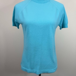 May include: A light blue short-sleeved sweater with a mock neck. The sweater is displayed on a mannequin. The sleeves are short and the knit fabric has a fine texture. The neckline and sleeves have a ribbed design.