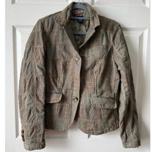 May include: A brown and tan plaid blazer with two front pockets and two buttons. The blazer has a notched collar and is hanging on a gray hanger. The interior lining has a floral pattern.