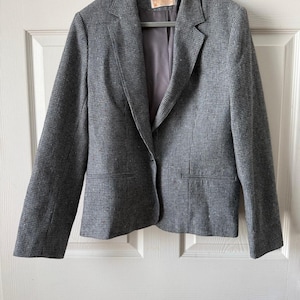 May include: A gray blazer with a subtle checkered pattern, hanging on a hanger. The blazer has a notched lapel, two front pockets, and a single-button closure. The interior lining is a solid gray color.