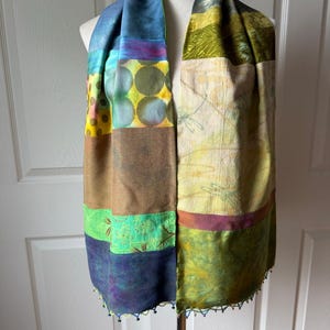 May include: A patchwork scarf featuring a variety of fabric panels in shades of blue, green, yellow, and brown. The scarf displays diverse patterns including circles, botanical prints, and solid textures, finished with small beaded fringe along the bottom edge.