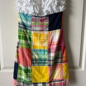 May include: A strapless dress with a patchwork design in various colors, including pink, yellow, green, and blue. The top of the dress features a white ruffled fabric. The dress is hanging on a white hanger.