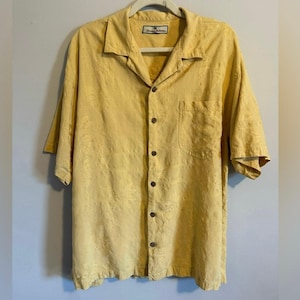 May include: Yellow button-down shirt with short sleeves and a chest pocket. The shirt is made of a textured fabric and has a relaxed fit. The shirt is hanging on a white hanger.