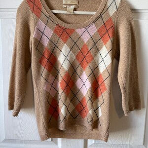May include: Tan sweater with a scoop neck and three-quarter sleeves. The sweater features a classic argyle pattern in shades of pink, orange, white, and black. The sweater is hanging on a white hanger against a white door.