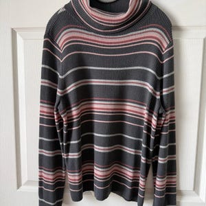 May include: A long-sleeved turtleneck sweater in a ribbed knit fabric. The sweater features horizontal stripes in gray, white, and shades of pink. The turtleneck is folded over. The sweater is hanging on a gray hanger.
