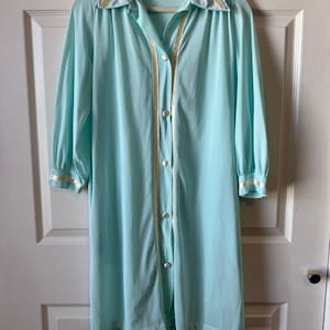 May include: A light blue vintage robe with a collared neckline and long sleeves. The robe features a button-down front with cream-colored trim and matching trim on the cuffs. The robe is hanging on a wooden hanger.