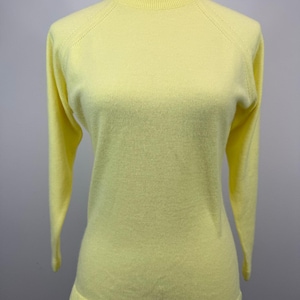 May include: A pale yellow sweater with a mock neck and long sleeves. The sweater is displayed on a mannequin. The sleeves have a raglan design. The sweater has a ribbed neckline and hem.