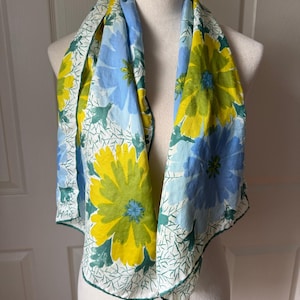 May include: A square scarf featuring a floral pattern with large yellow and light blue flowers on a white background. The design includes green foliage accents and thin, dark green branch-like lines. The scarf has a dark green hem and the name Vera is printed in the corner.