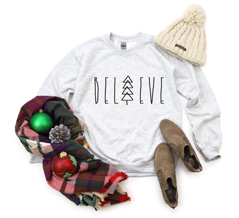 Puede incluir: Una sudadera gris con las palabras "BELIEVE" impresas en la parte delantera, con un &aacute;rbol de Navidad estilizado en lugar de la "I". La sudadera est&aacute; colocada sobre un fondo blanco con una bufanda de cuadros, dos adornos navide&ntilde;os, una pi&ntilde;a y una boina blanca.