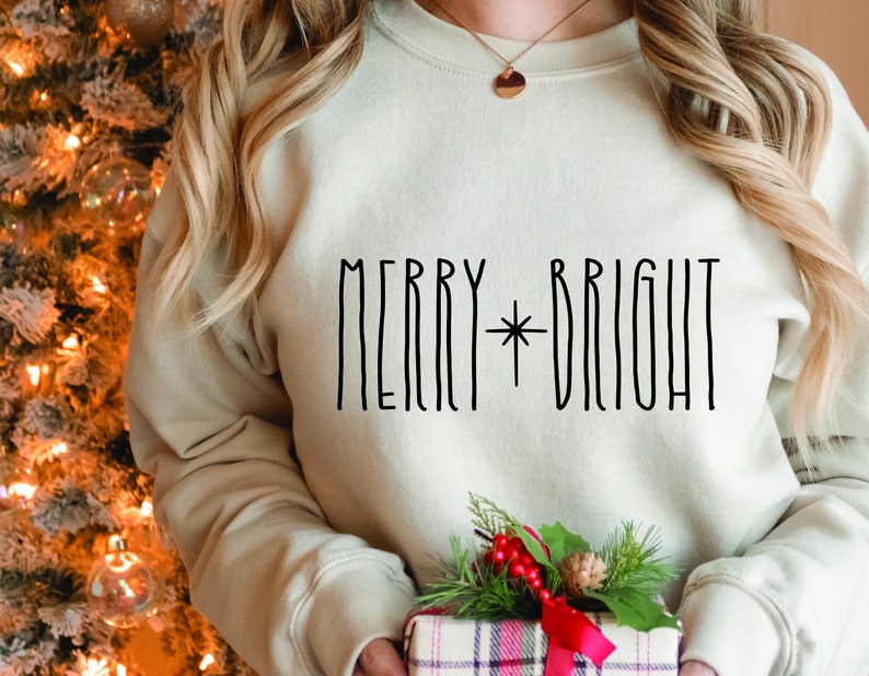 Puede incluir: Una sudadera blanca con el texto "Merry*Bright" en letras negras. La sudadera est&aacute; siendo sostenida por una persona frente a un &aacute;rbol de Navidad.