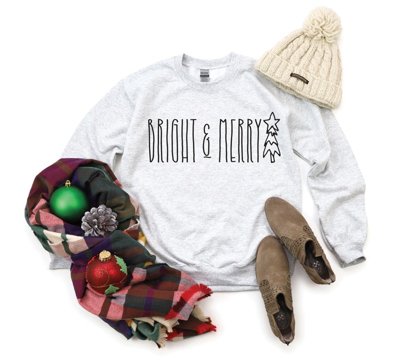 Puede incluir: Una sudadera gris con el texto "BRIGHT & MERRY" y un gr&aacute;fico de &aacute;rbol de Navidad estilizado. La sudadera est&aacute; colocada sobre un fondo blanco con una bufanda de cuadros, una bola de Navidad verde y roja, una pi&ntilde;a y un gorro beige.