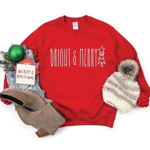 Puede incluir: Sudadera roja de Navidad con el texto "Bright & Merry" en blanco con un gr&aacute;fico de &aacute;rbol de Navidad estilizado. La sudadera est&aacute; colocada sobre un fondo blanco con un par de botas marrones, unos vaqueros azules, un adorno verde y un gorro blanco de punto con un pomp&oacute;n de piel sint&eacute;tica.