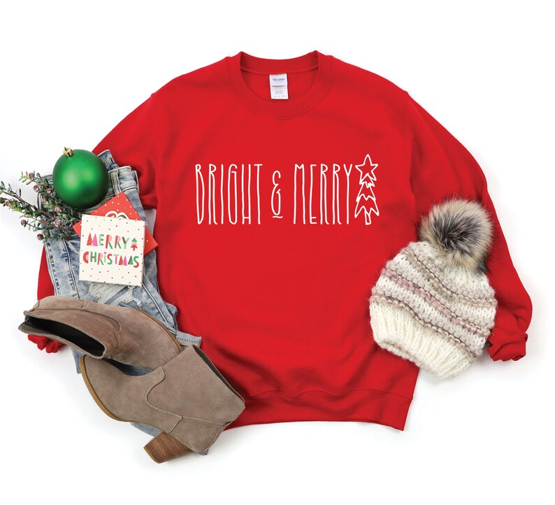 Puede incluir: Sudadera roja de Navidad con el texto "Bright & Merry" en blanco con un gr&aacute;fico de &aacute;rbol de Navidad estilizado. La sudadera est&aacute; colocada sobre un fondo blanco con un par de botas marrones, unos vaqueros azules, un adorno verde y un gorro blanco de punto con un pomp&oacute;n de piel sint&eacute;tica.