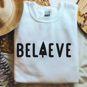 Op de afbeelding: Witte trui met het woord "BELIEVE" in zwart gedrukt, met een klein hartje in een gestileerde boom ter vervanging van de "I".