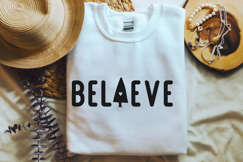 Op de afbeelding: Witte trui met het woord "BELIEVE" in zwart gedrukt, met een klein hartje in een gestileerde boom ter vervanging van de "I".