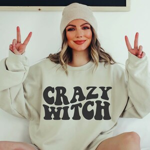 May include: A light beige sweatshirt with the text "CRAZY WITCH" in black, retro-style lettering. The model is wearing the sweatshirt and a beige beanie.
