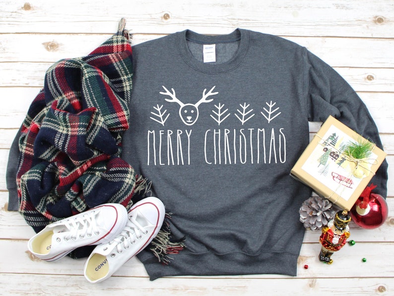 Puede incluir: Una sudadera gris con un gr&aacute;fico blanco de reno y &aacute;rboles de Navidad y el texto "Merry Christmas". La sudadera est&aacute; colocada sobre una superficie de madera blanca con una bufanda de cuadros, zapatillas blancas, una peque&ntilde;a caja de regalo, un cascanueces y una bola de Navidad roja.