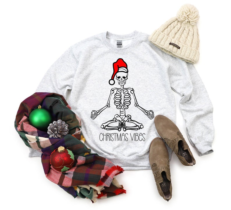 Puede incluir: Una sudadera gris con un esqueleto que lleva un gorro de Pap&aacute; Noel y est&aacute; sentado en una postura de yoga. El texto "Christmas Vibes" est&aacute; impreso debajo del esqueleto. La sudadera est&aacute; colocada sobre un fondo blanco con una bufanda de cuadros, un adorno navide&ntilde;o verde, una pi&ntilde;a y un par de botas marrones.