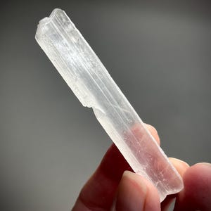Puede incluir: Un cristal claro y alargado con una textura ligeramente rugosa. El cristal se sostiene en una mano, mostrando su cualidad translúcida. El cristal tiene forma rectangular, con estrías internas visibles. El fondo es de un gris suave.