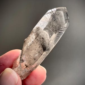 Può includere: Un cristallo di quarzo trasparente con un'inclusione grigia e nuvolosa. Il cristallo è appuntito a un'estremità e ha una superficie ruvida e naturale.
