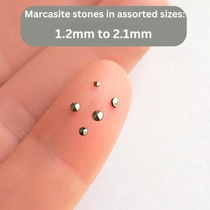 Puede incluir: Primer plano de pequeñas piedras de marcasita facetadas en varios tamaños, que van de 1,2 mm a 2,1 mm. Las piedras son de un color oscuro y metálico y se muestran en un dedo. La imagen incluye el texto "Bead Dream Shop".