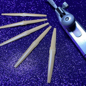 May include: Five wooden oboe reeds arranged in a fan shape on a blue sparkly background. A silver oboe reed tool with the word "KOAS" printed on it is in the upper right corner of the image.