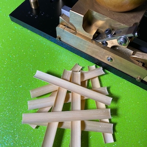 Puede incluir: Primer plano de un dispositivo mecánico con componentes de latón y negros, y una esfera de madera. Delgadas tiras de madera de color claro están apiladas sobre una superficie verde brillante y brillante. El dispositivo parece ser una herramienta para trabajar la madera.