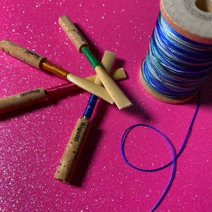 May include: Close-up of reeds for a musical instrument, with cork and colorful thread accents. The reeds are labeled "Rigotti". A spool of blue, green, and white thread sits nearby, with a length of blue thread extending from it. The background is a bright pink, glittery surface.