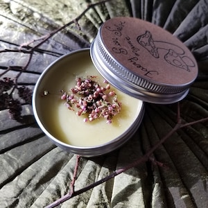 May include: A small, open metal tin filled with a pale yellow balm, topped with small pink flowers. The lid, with a mushroom illustration and text, sits beside it. The background is a textured, olive green fabric with twigs.