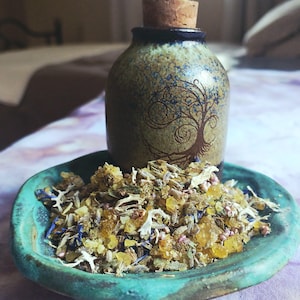 May include: A ceramic jar with a cork stopper, featuring a tree of life design, sits above a teal-colored ceramic dish filled with dried herbs and crystals. The jar is approximately 4 inches tall.