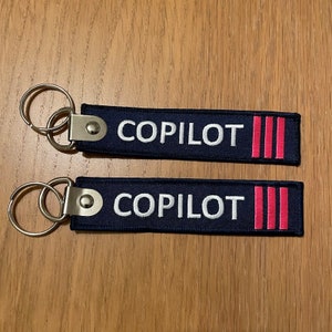 May include: Two blue keychains with silver key rings. Each keychain has white embroidered text that says "COPILOT" and three pink stripes.