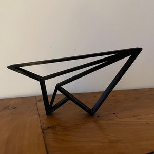 Könnte beinhalten: Schwarze Papierflieger-Skulptur aus Metall. Die Skulptur besteht aus dünnem Metall und ist in Form eines Papierfliegers gestaltet. Sie steht auf einer Holzoberfläche.