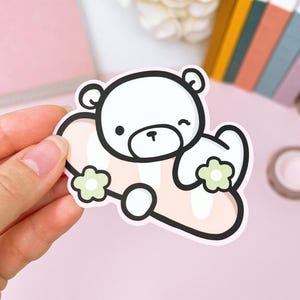 Op de afbeelding: Een witte berensticker met een zwarte omtrek, knipogend en met twee lichtgroene bloemen. De beer staat op een roze hartvormig ontwerp met een zwarte omtrek. De sticker wordt door een hand tegen een roze achtergrond gehouden.