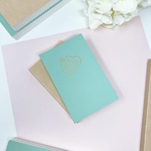 May include: Two notebooks, one teal and one brown, with a gold design of an open book with stars on the teal notebook.