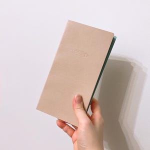 May include: A light pink notebook with a subtle embossed pencil design. The notebook has a light green spine.