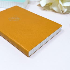 May include: A small, mustard yellow leather-bound notebook with a white leaf design on the cover. The notebook has white pages and a white spine.