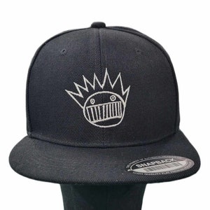 May include: Black baseball cap with a white embroidered graphic of a cartoon face with a crown and teeth. The cap has a black adjustable strap with a white "SNAPBACK" label.