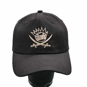 Pode incluir: Boné de beisebol preto com um logotipo branco bordado de um rosto estilizado com uma coroa e espadas cruzadas.