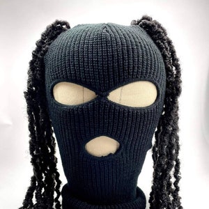May include: A black knitted balaclava with eye and mouth openings. The balaclava has long, black, curly faux hair extensions attached on each side. The hat is made of a thick, textured knit material, offering a distinctive and fashionable appearance.