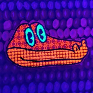 May include: An orange and black alligator patch with a grid pattern and star details. The alligator has teal eyes and is set against a dark blue background with a repeating pattern of purple ovals.