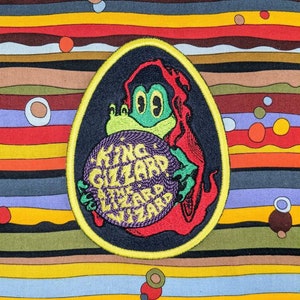 May include: A yellow, green, red, and purple embroidered patch with a cartoon frog holding a purple orb. The orb has the text "KING GIZZARD AND THE LIZARD WIZARD" written on it. The patch is on a striped background.