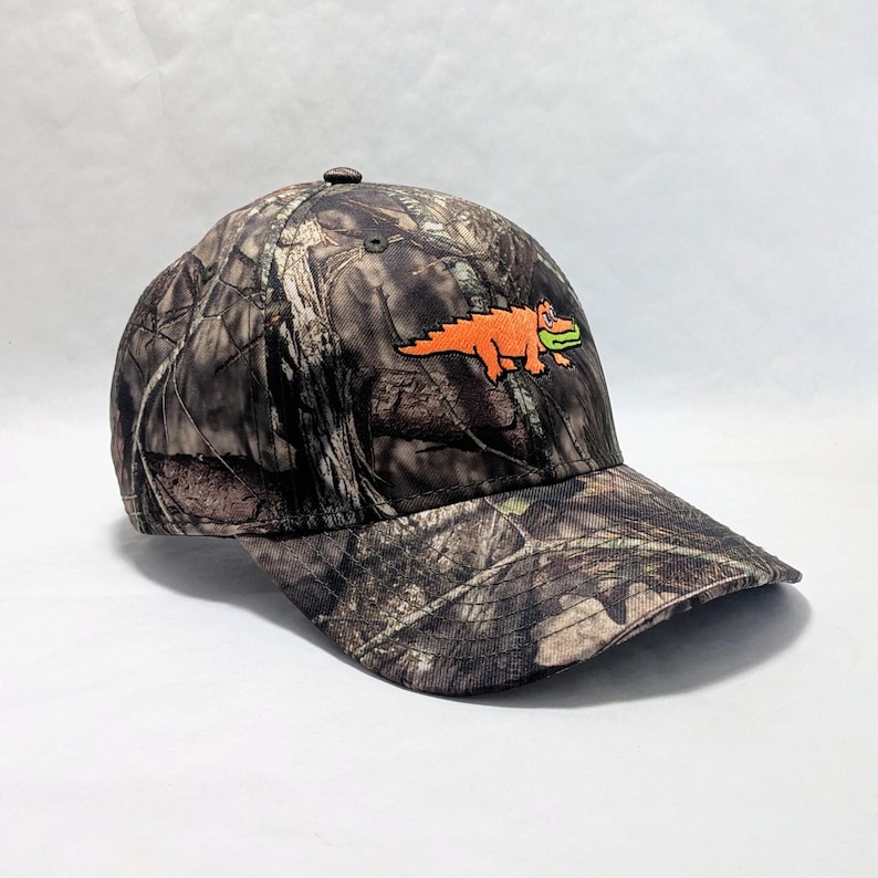 Op de afbeelding: Camouflage baseballpet met een geborduurde oranje alligator en groene details. De pet heeft een gebogen klep en een knoop aan de bovenkant. Het patroon is een mix van bruin, groen en beige, dat op een bos lijkt.