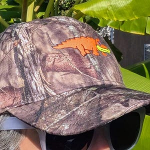 Op de afbeelding: Camouflage baseballpet met een oranje dinosaurusafbeelding. De pet heeft een bruin, groen en beige camouflagepatroon. De dinosaurus houdt een geel voorwerp vast. De pet heeft een gebogen klep.