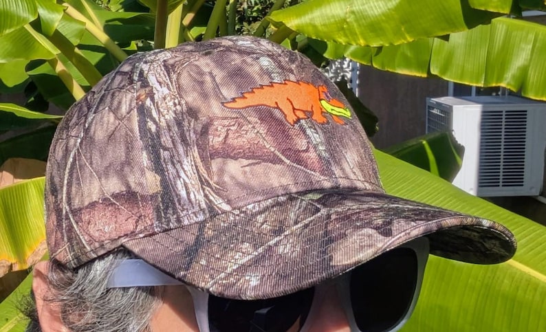 Op de afbeelding: Camouflage baseballpet met een oranje dinosaurusafbeelding. De pet heeft een bruin, groen en beige camouflagepatroon. De dinosaurus houdt een geel voorwerp vast. De pet heeft een gebogen klep.