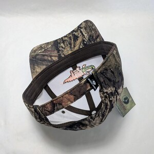 Op de afbeelding: Een camouflage baseballpet met een bruine rand en een witte binnenkant. De binnenkant heeft een geborduurd ontwerp van een cartoon alligator met een hengel. De pet heeft een Mossy Oak label.