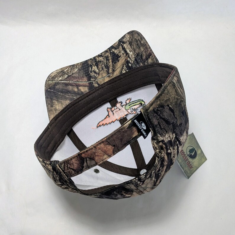 Op de afbeelding: Een camouflage baseballpet met een bruine rand en een witte binnenkant. De binnenkant heeft een geborduurd ontwerp van een cartoon alligator met een hengel. De pet heeft een Mossy Oak label.